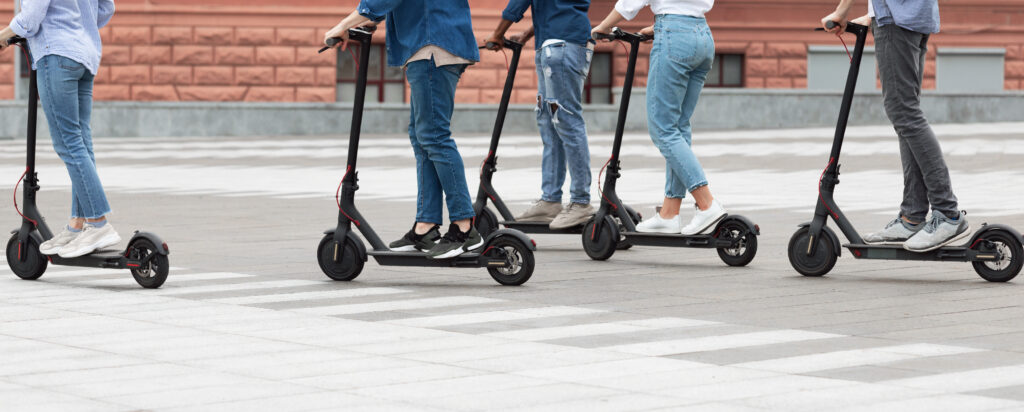 Five people ride electric scooters outdoors on a paved area. Only their legs and lower bodies are visible, and they are wearing casual clothes and sneakers. The background includes a brick building wall.