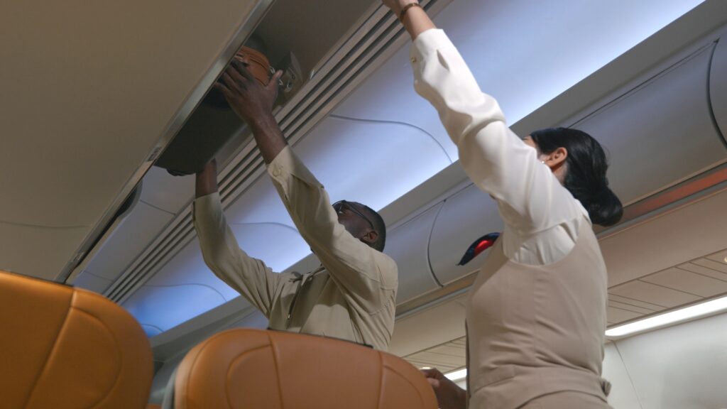 A flight attendant assists a passenger in placing his bag into an overhead compartment on an airplane, with brown leather seats visible in the foreground.