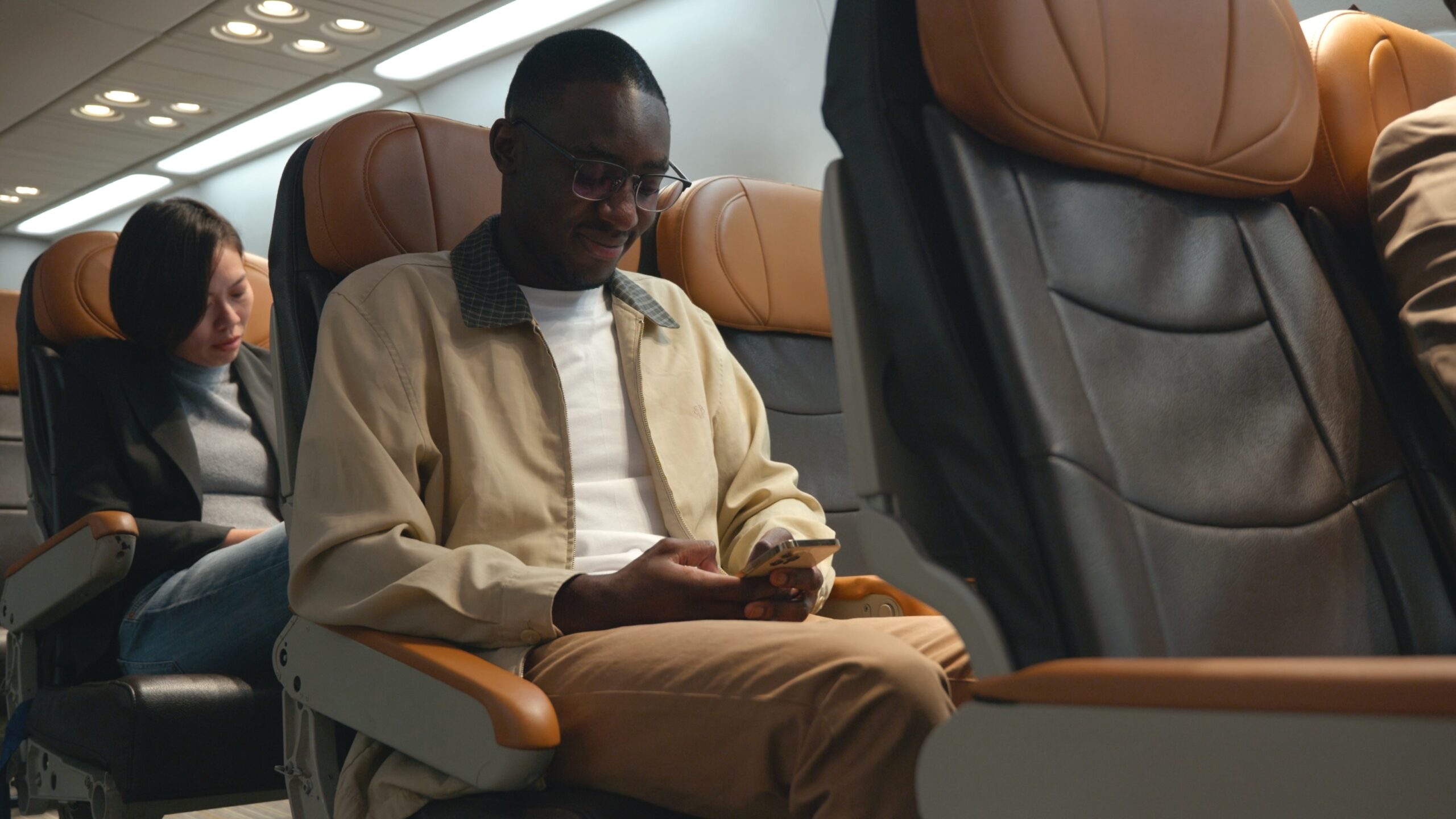 A man sits in an airplane seat, smiling and looking at his phone. Another passenger sits behind him, reading. The cabin has brown and black leather seats and overhead lights are on.