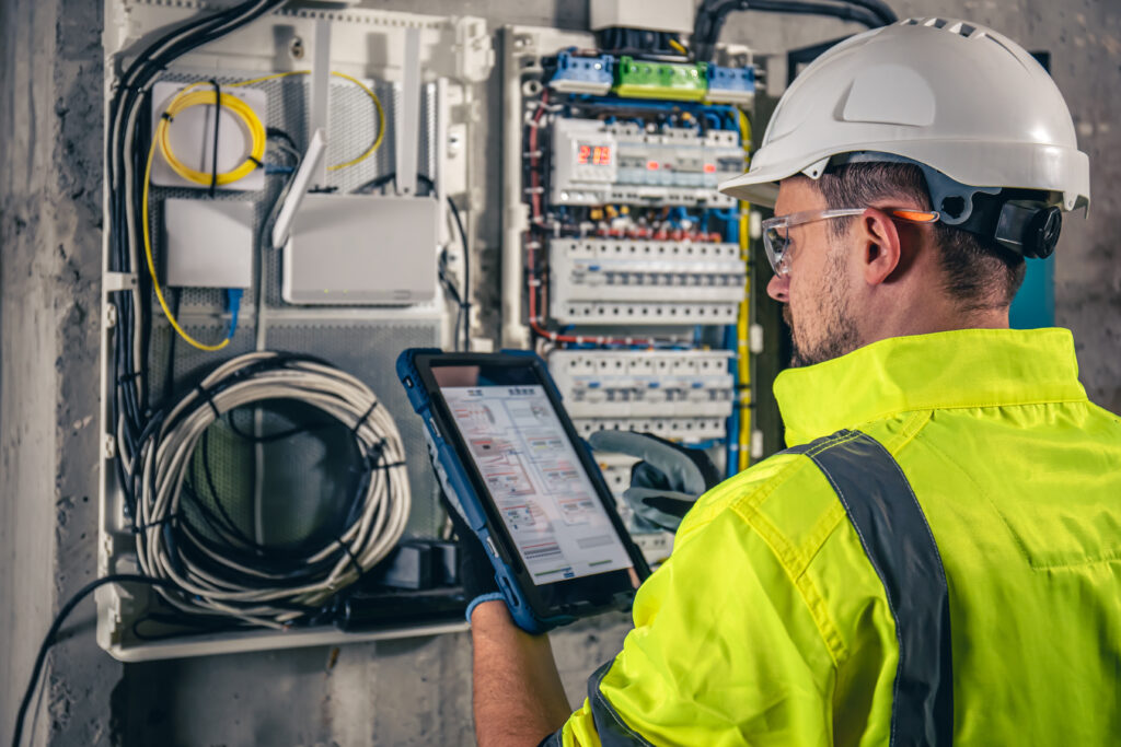 A worker in a hard hat and high-visibility jacket uses a tablet to inspect or monitor electrical control panels and wiring on a wall, likely performing maintenance or troubleshooting.