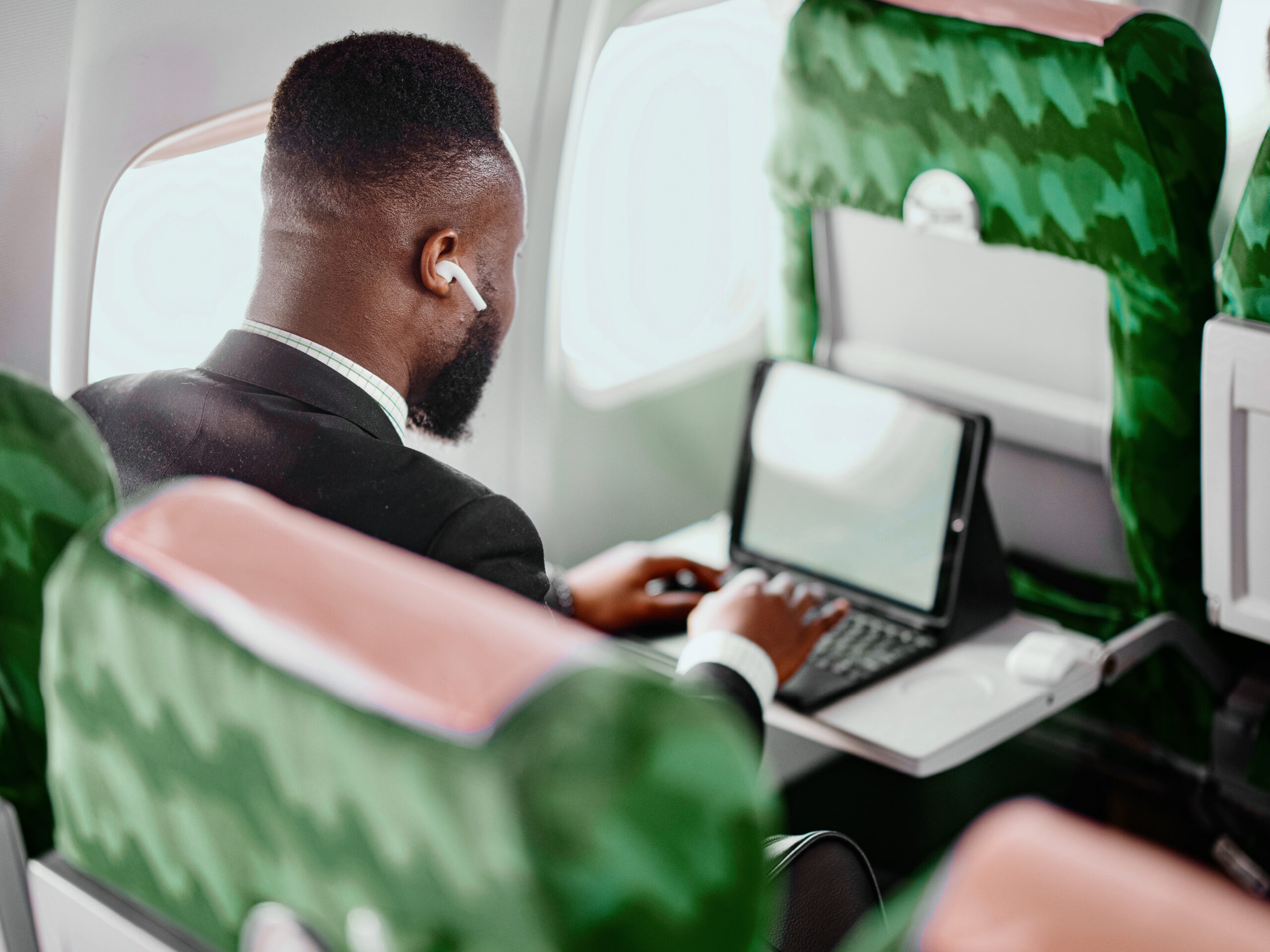 A man in a suit, wearing wireless earbuds, works on a laptop at his seat on an airplane.