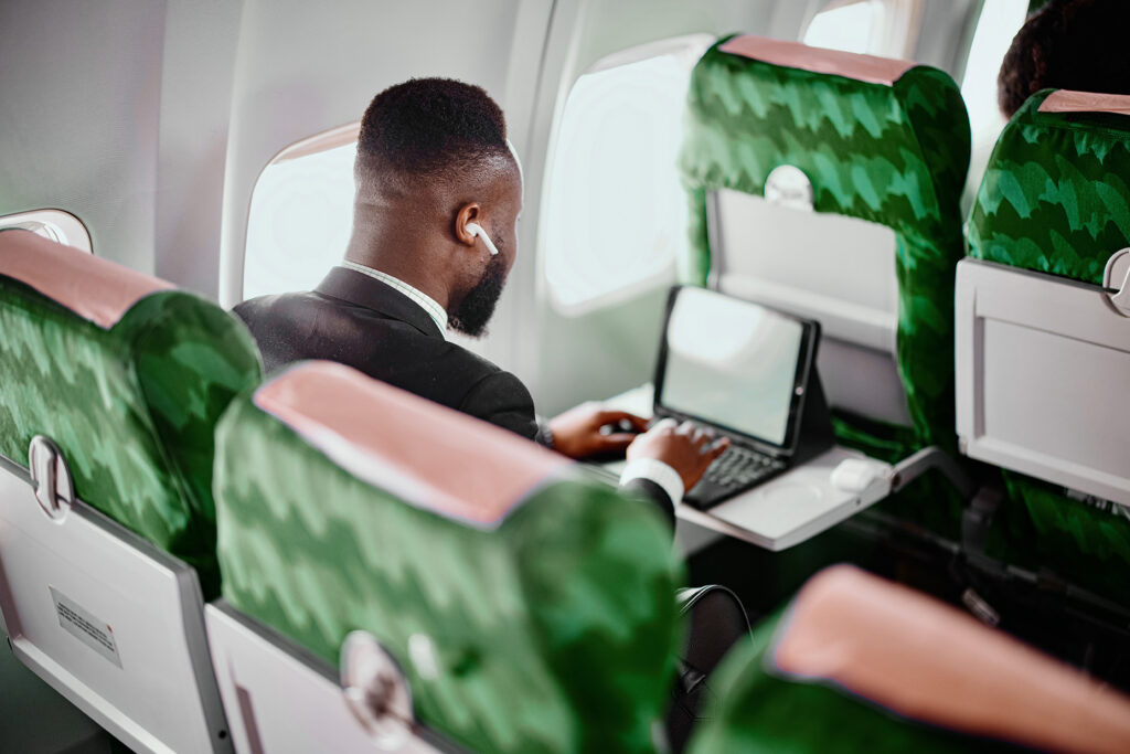 A man in a suit works on a laptop while sitting in an airplane seat with green headrests. He is wearing earbuds and facing an airplane window.