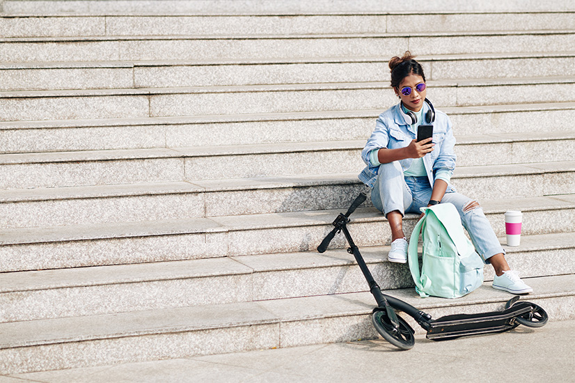 A young woman in casual clothes and sunglasses sits on outdoor steps, looking at her phone. She has a mint backpack and takeaway coffee beside her, with an electric scooter lying on the steps in front.