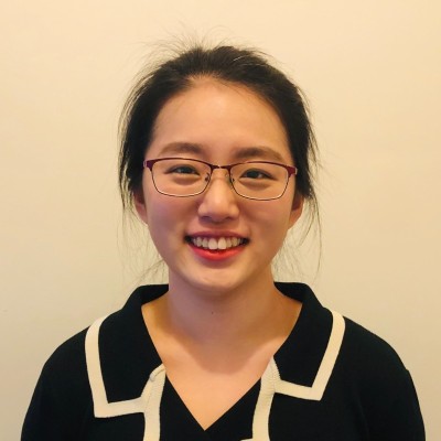 A young woman with glasses and dark hair tied back smiles at the camera. She is wearing a black top with a white-trimmed collar, and is standing in front of a plain, light-colored background.
