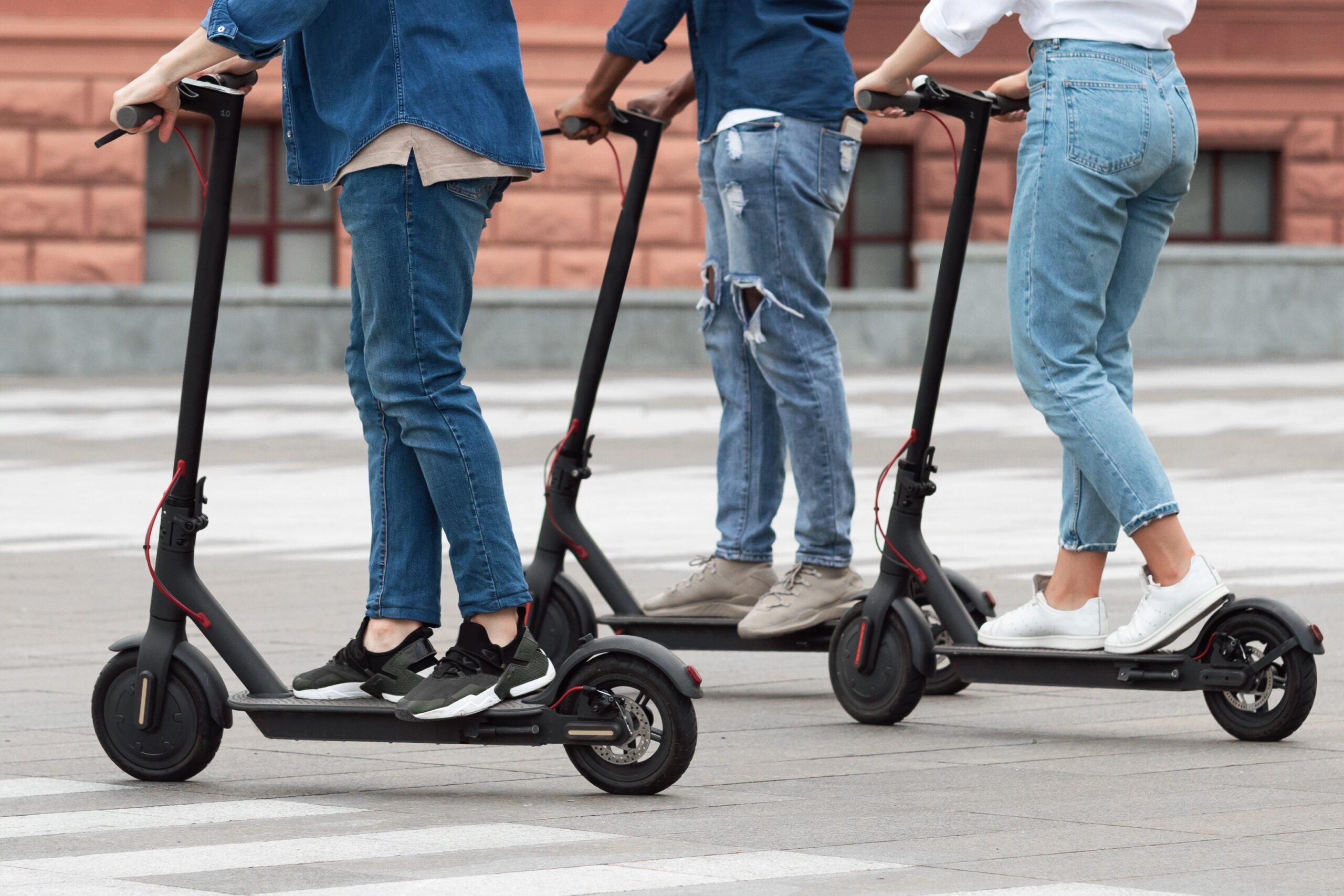 Three people wearing jeans and sneakers ride electric scooters on a city street, with a building featuring red bricks and windows in the background. The photo is taken from the waist down.