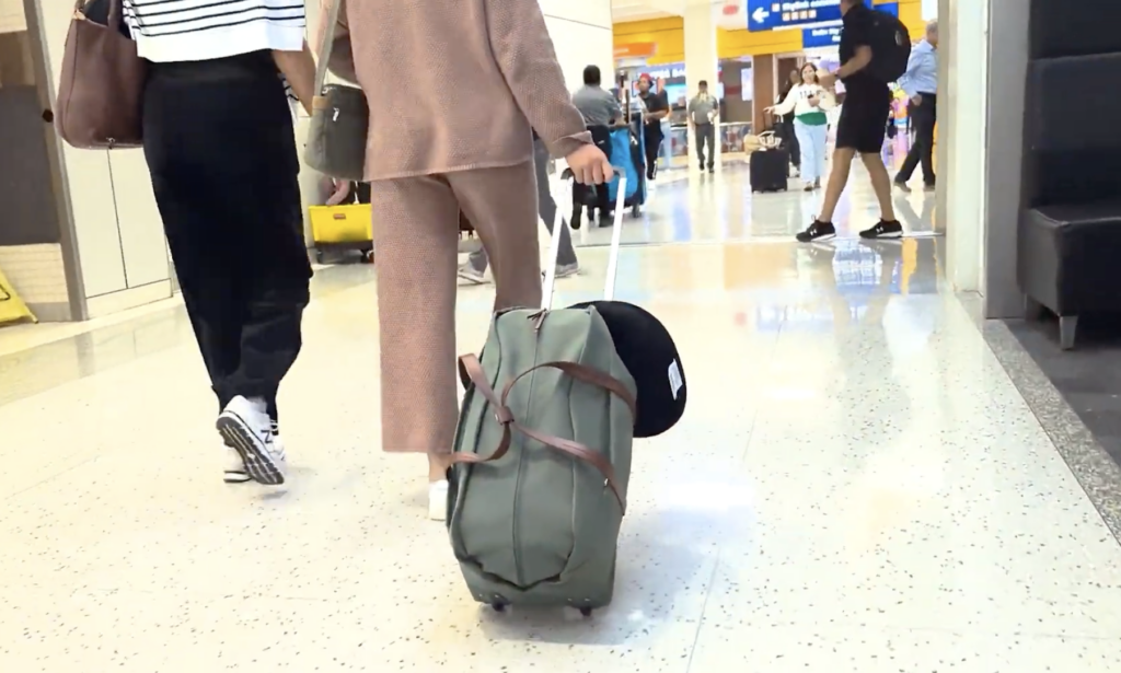 A person in a pink outfit pulls a green wheeled suitcase through a busy airport terminal, surrounded by other travelers.