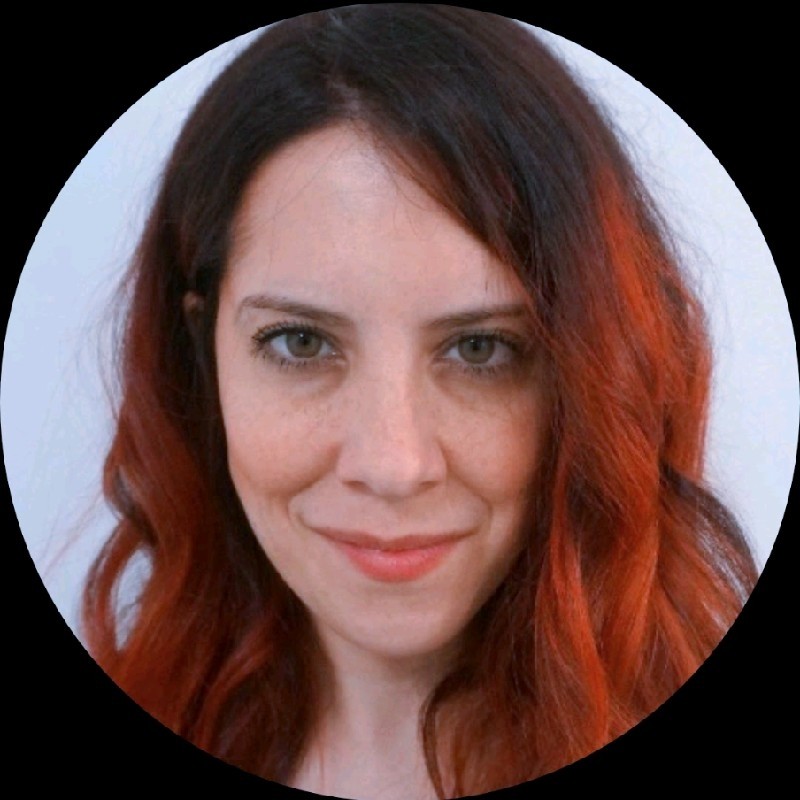 A woman with long, wavy, dark red hair and fair skin looks directly at the camera, smiling slightly. The background is plain white, and the image is cropped in a circular frame.