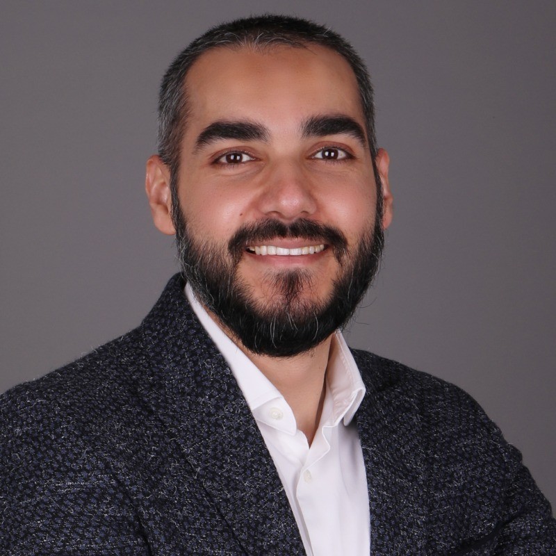 A man with short dark hair and a full beard smiles at the camera. He is wearing a textured dark blazer over a white shirt and is posed against a plain gray background.