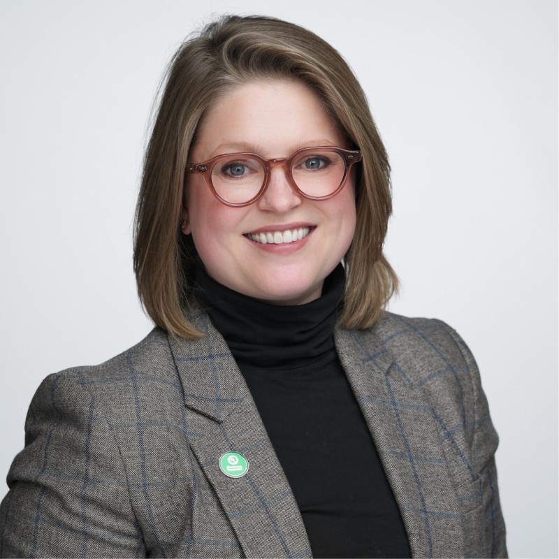 A woman with short, light brown hair and round glasses smiles at the camera. She is wearing a plaid blazer over a black turtleneck and a green circular pin on her lapel. The background is plain and light-colored.
