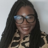 A woman with long braids, wearing large black glasses and a beige and black patterned top, smiles at the camera against a plain light background.