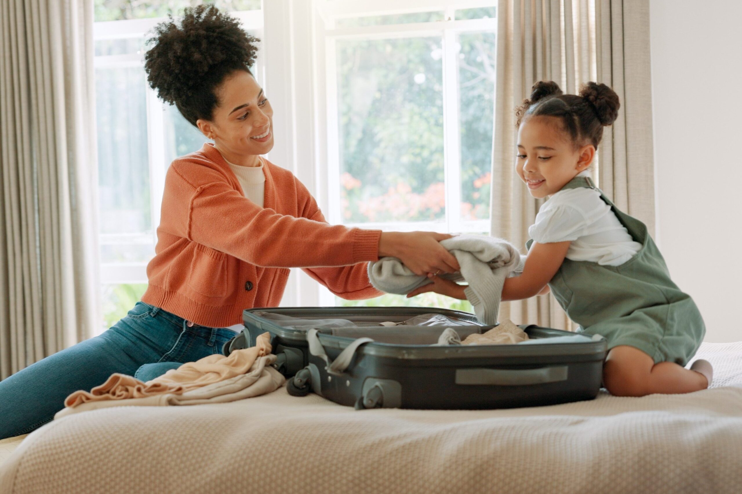 A woman and a young girl smile while packing clothes into an open suitcase on a bed, with sunlight streaming through windows behind them.
