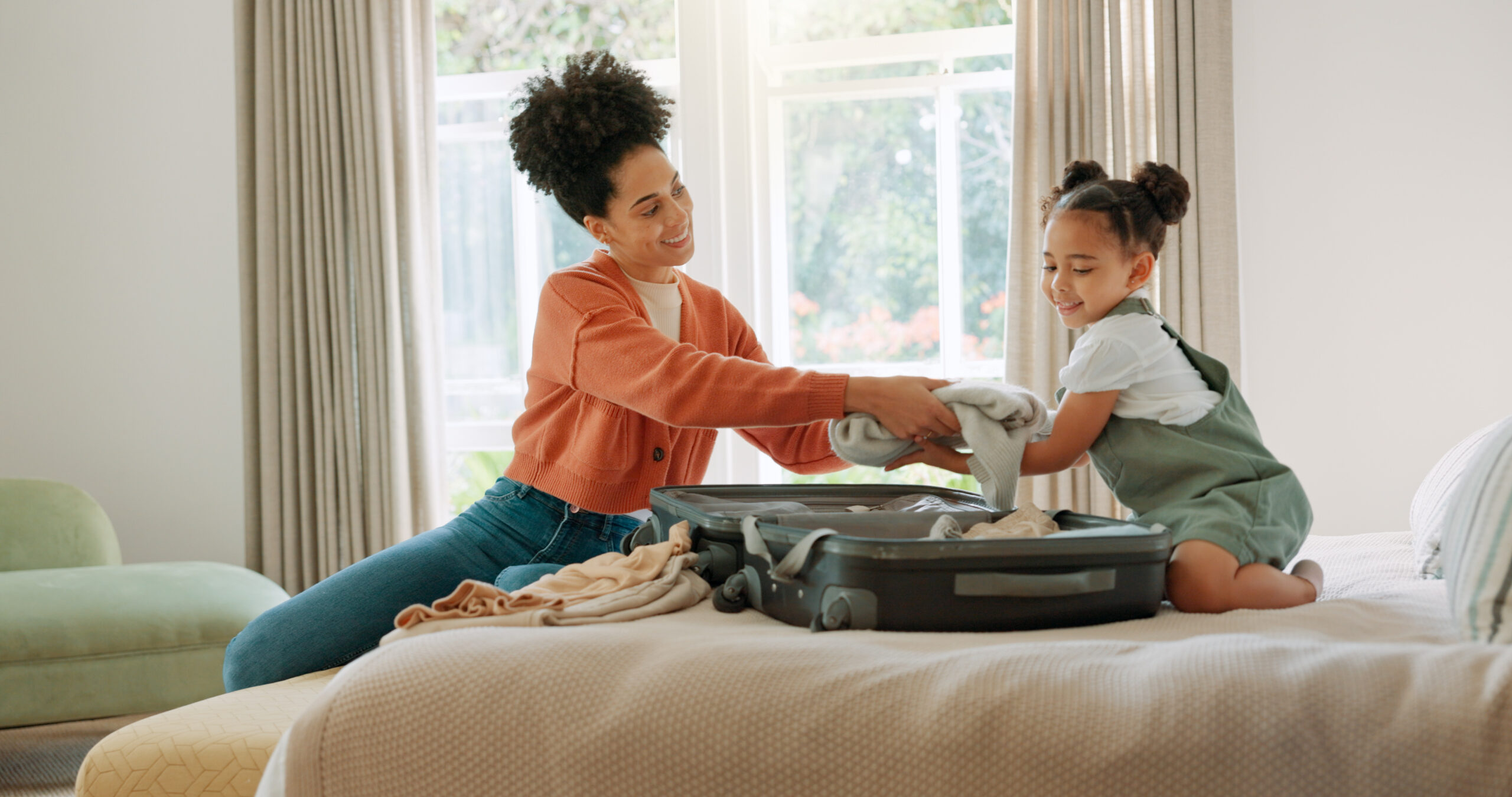 A woman and a young girl sit on a bed, smiling and packing clothes into an open suitcase together, with large windows in the background letting in natural light.