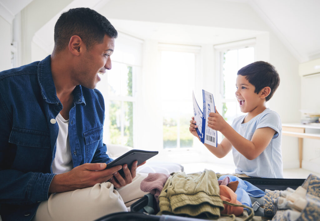 A man and a child smile at each other while packing a suitcase. The man holds a tablet, and the child excitedly holds travel documents and a boarding pass in a bright, sunlit room.