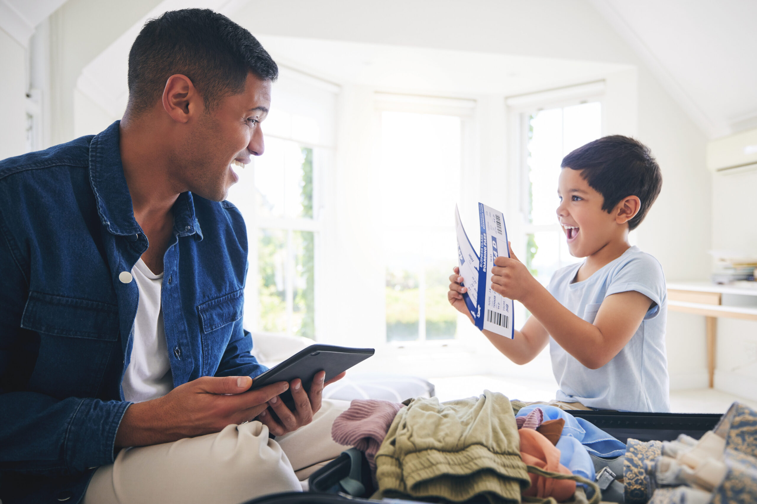A man and a child smile at each other while packing a suitcase. The man holds a tablet, and the child excitedly holds travel documents and a boarding pass in a bright, sunlit room.