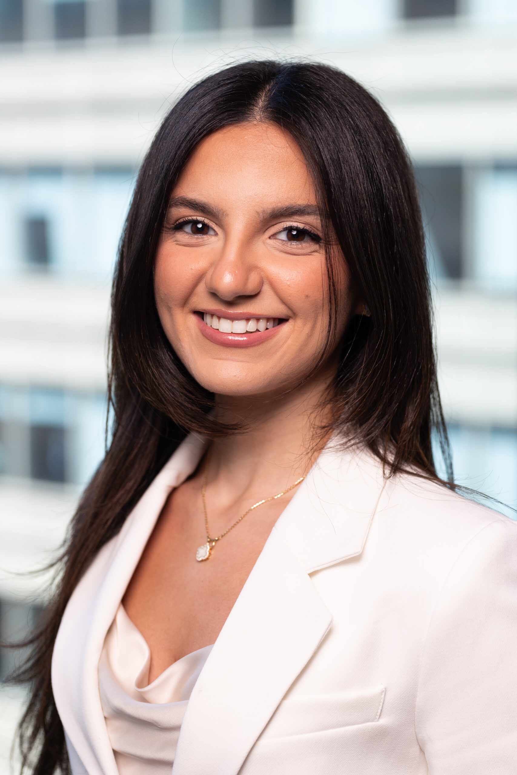 A woman with long dark hair, wearing a white blazer and a gold necklace, smiles at the camera. The background is blurred with white and blue horizontal lines, suggesting an office building.
