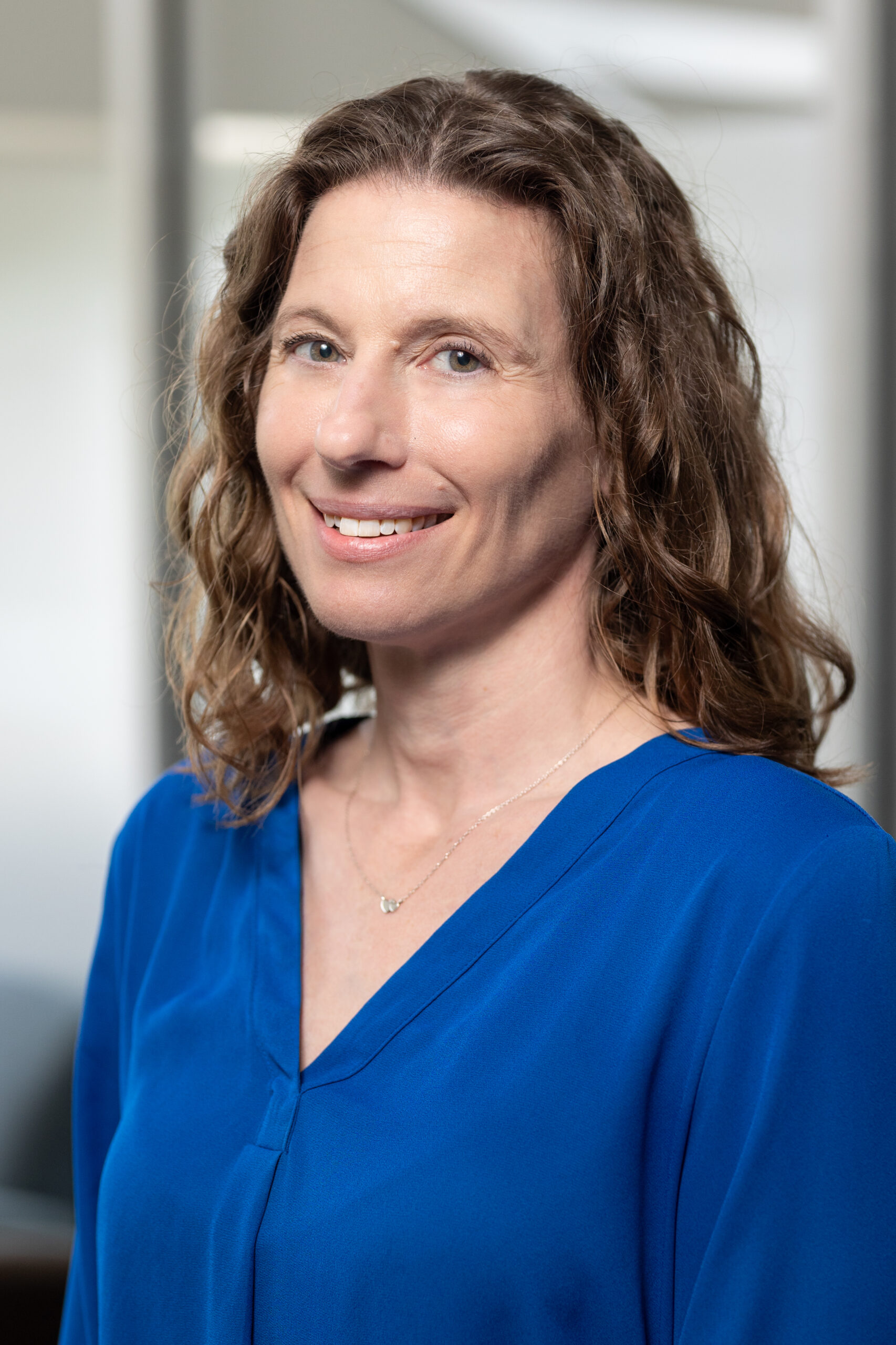 A woman with wavy, shoulder-length brown hair smiles at the camera. She is wearing a royal blue blouse and a delicate necklace. The background is softly blurred, suggesting an indoor setting.