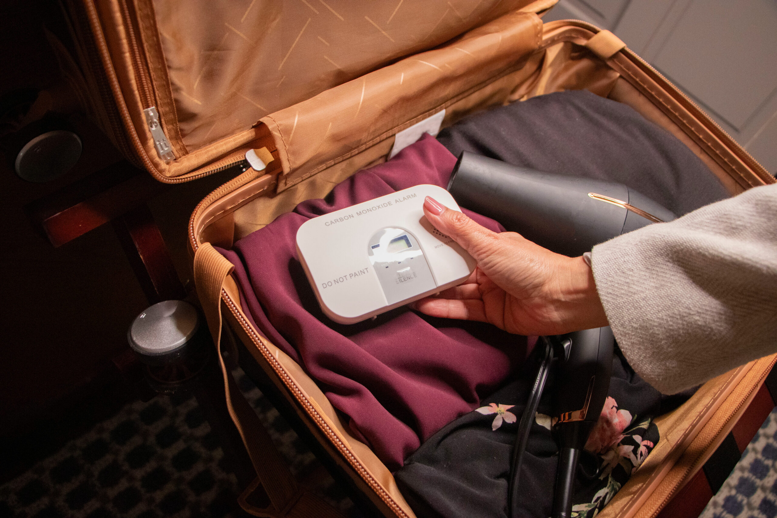 A person packs a suitcase, placing a white carbon monoxide alarm on top of folded clothes next to a black hair dryer inside the open luggage.