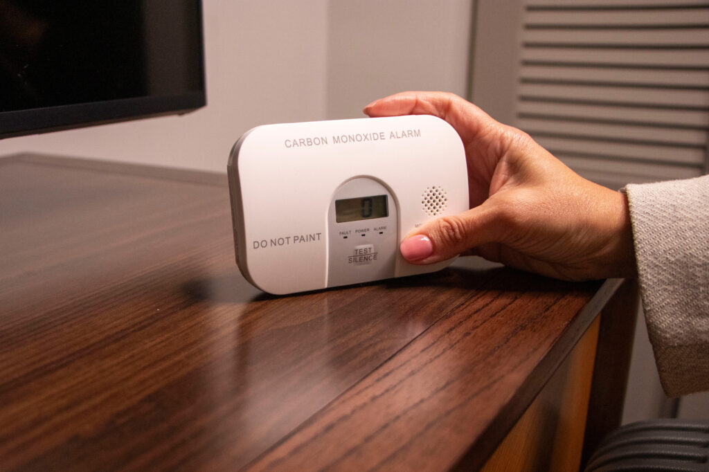 A hand holds a carbon monoxide alarm on a wooden table, displaying a reading of 0 on its digital screen. The device has “DO NOT PAINT” and “TEST WEEKLY” instructions on the front.