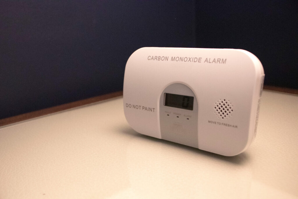 A carbon monoxide alarm with a digital display showing "0" sits on a light-colored surface against a dark wall. The device has labels "DO NOT PAINT" and "MOVE TO FRESH AIR.