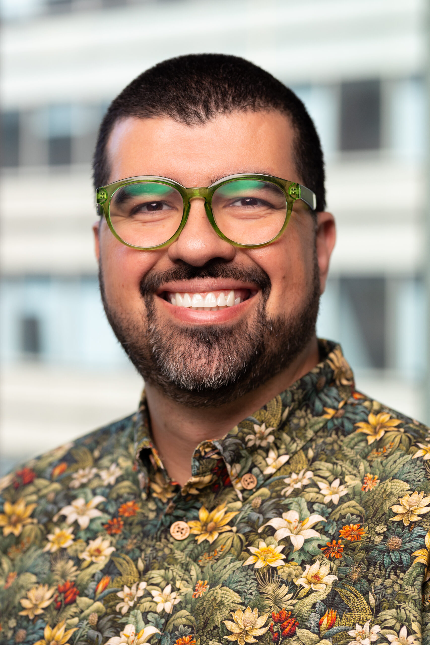 A smiling person with short dark hair, a beard, and green glasses is wearing a floral shirt. The background is softly blurred, showing a modern building with large windows.