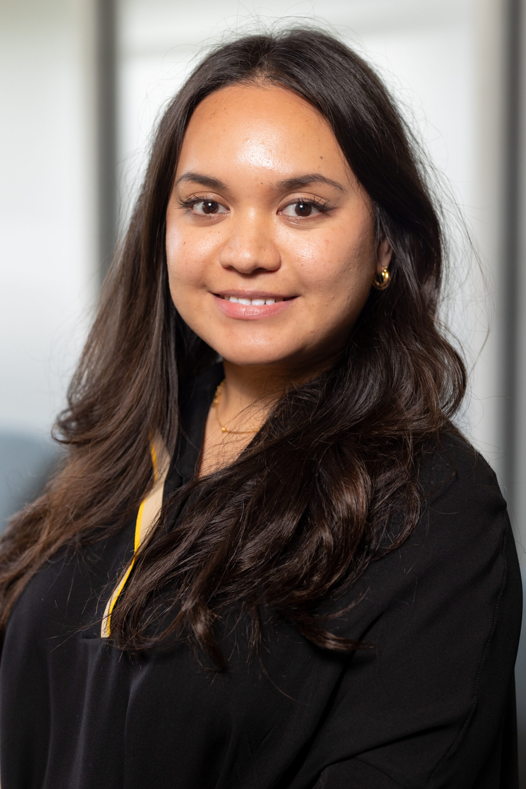A woman with long dark hair, wearing a black top with a yellow detail and gold earrings, smiles at the camera in a brightly lit indoor setting.