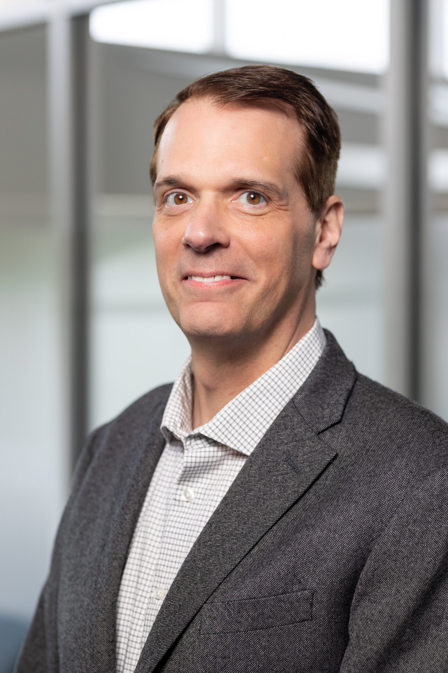 A man in a gray suit jacket and checked shirt stands in front of a blurred office background, looking at the camera with a neutral expression.