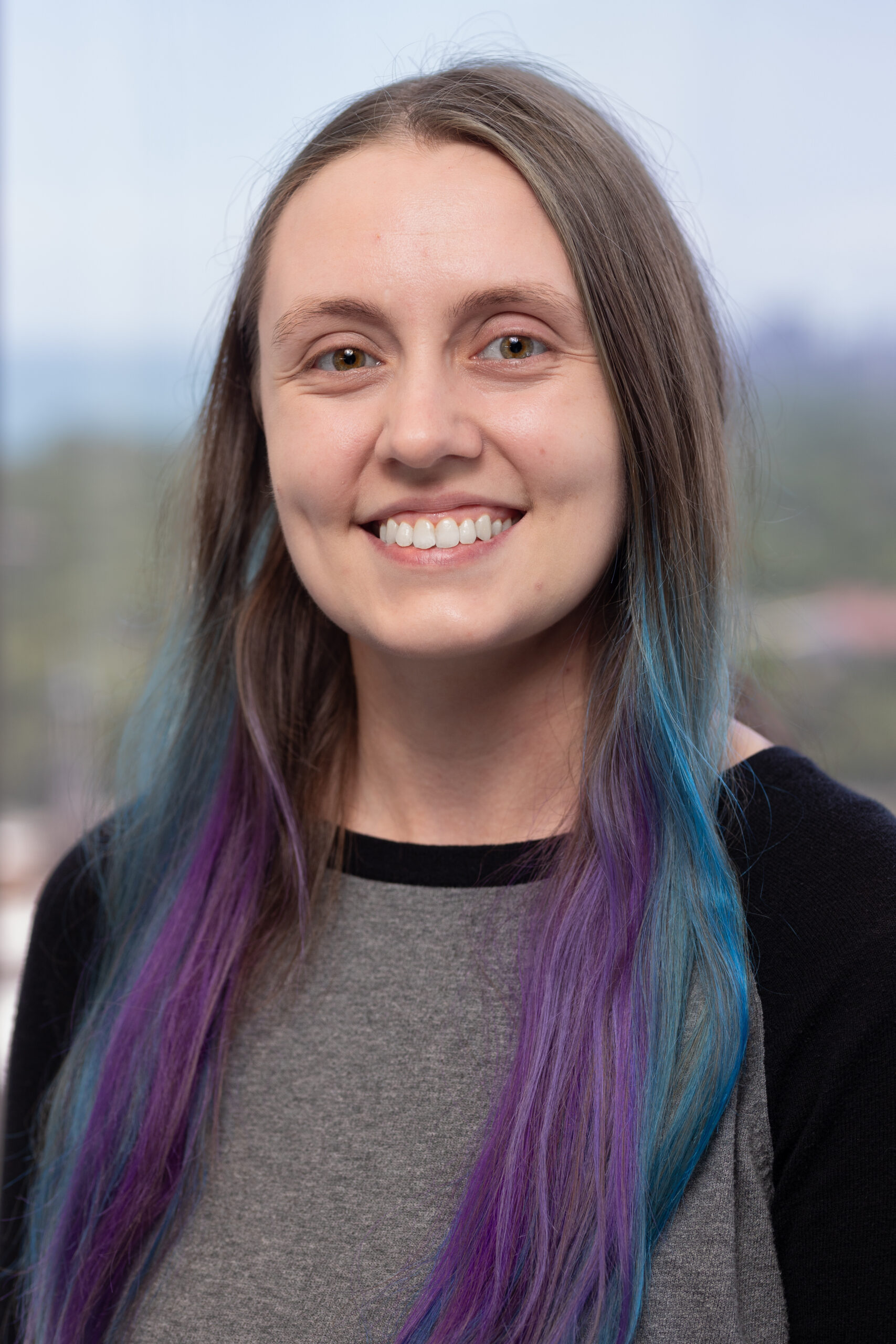 A young woman with long brown hair featuring purple and blue tips smiles at the camera. She is wearing a grey and black shirt, and the background is softly blurred.