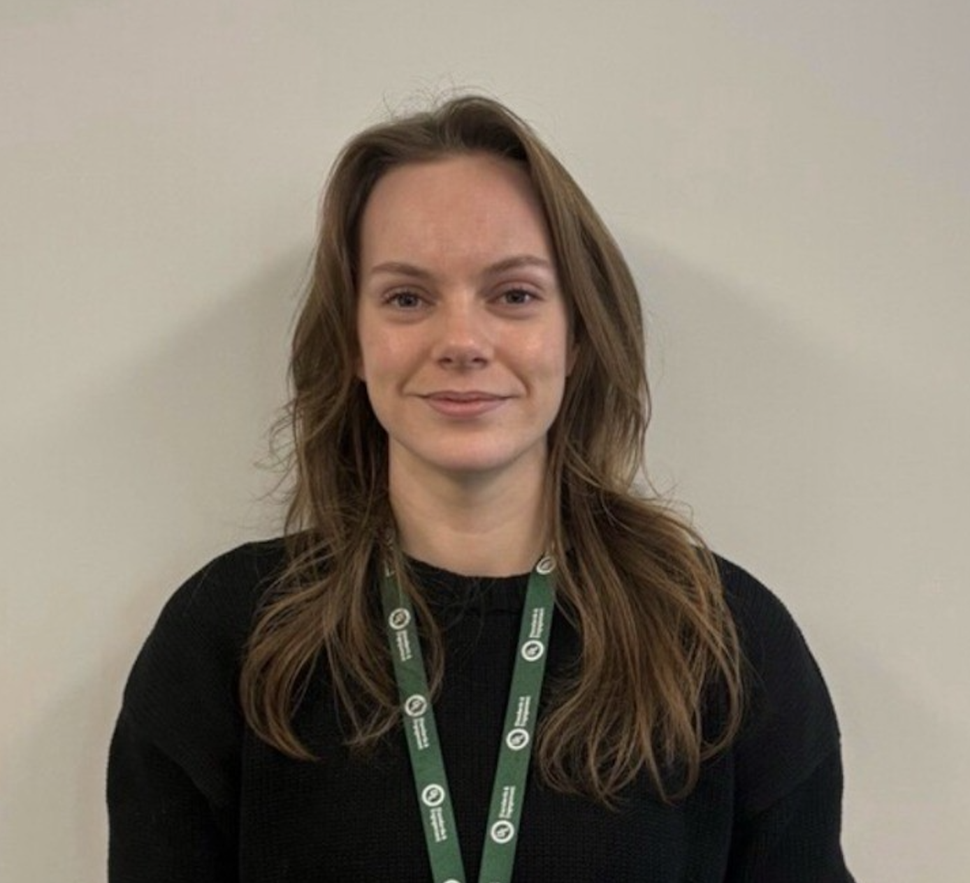 A woman with long brown hair wearing a black sweater and a green lanyard stands in front of a plain light-colored wall, smiling slightly at the camera.