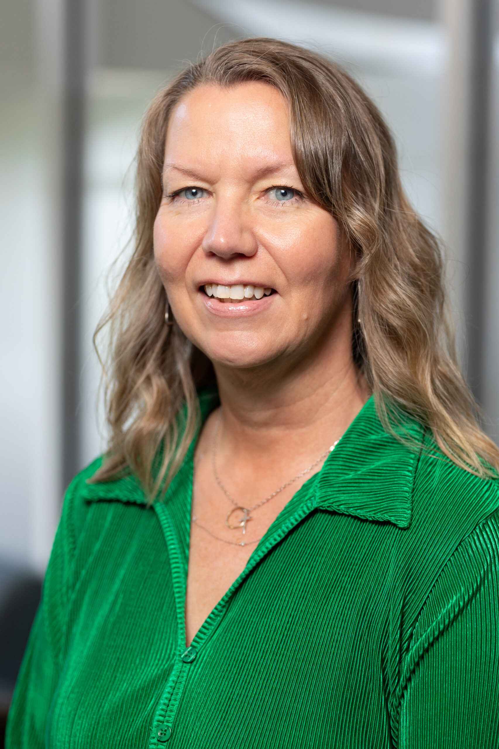 A woman with wavy blonde hair smiles at the camera. She is wearing a bright green collared shirt and a delicate necklace. The background is softly blurred, suggesting an indoor setting.