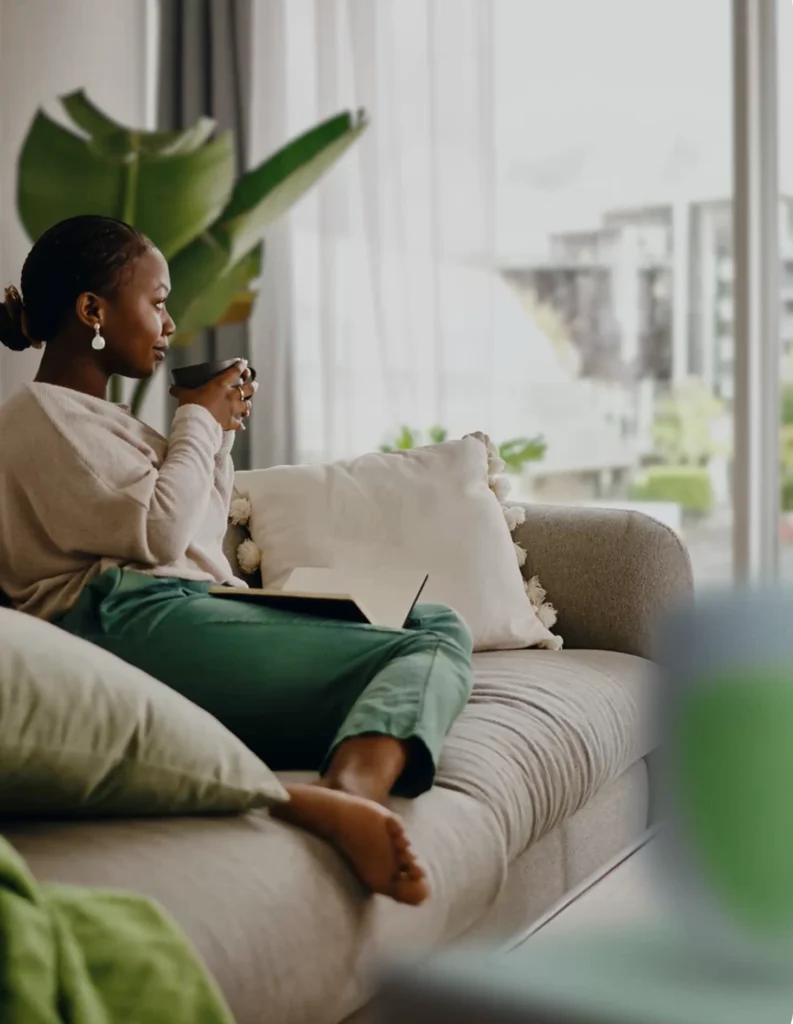 A woman sits on a sofa with her legs crossed, holding a mug and looking out of a large window. She is surrounded by green plants and cushions, her notebook resting on her lap—creating a calm, cozy atmosphere in line with UL Standards & Engagement.