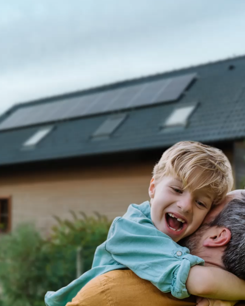 A smiling child with blond hair embraces an adult outdoors, in front of a house with solar panels on the roof that meet UL Standards & Engagement for safety. The child appears happy and joyful.