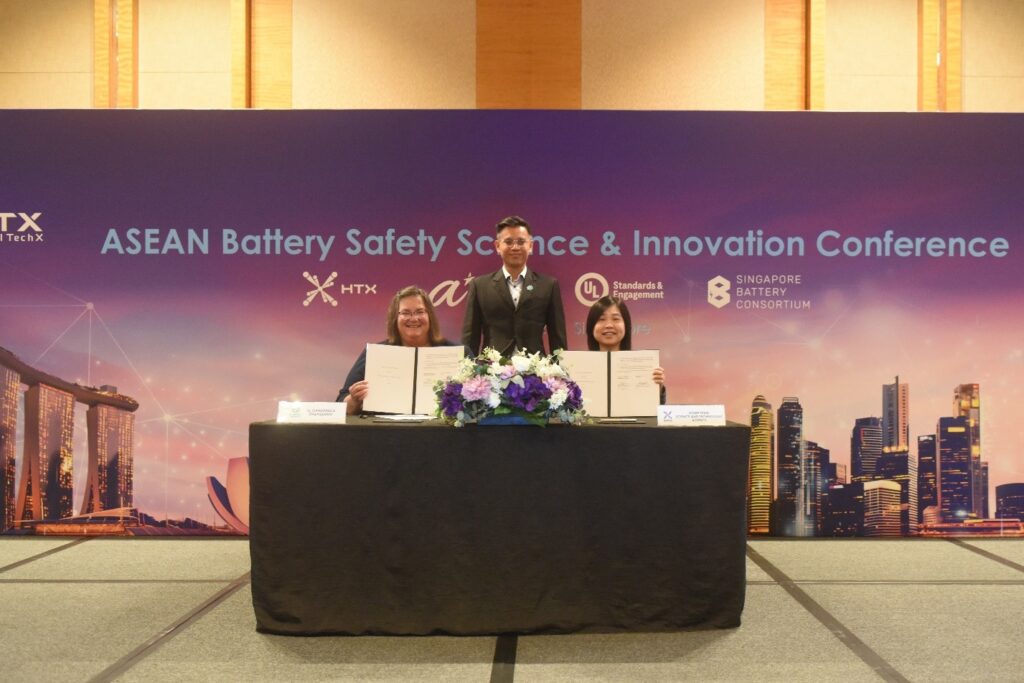 Three people pose at a table with documents during the ASEAN Battery Safety Science & Innovation Conference. The backdrop features event and sponsor logos, with a cityscape graphic.