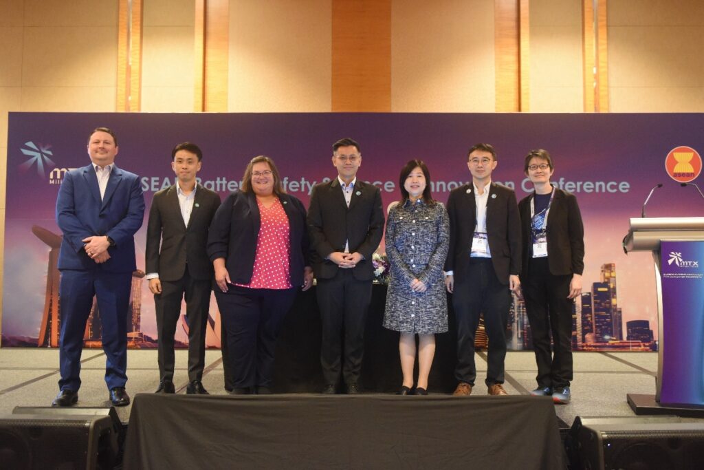 Seven people in formal attire stand on a stage at a conference. A podium is on the right, and a screen behind them displays conference branding with cityscape imagery and logos.