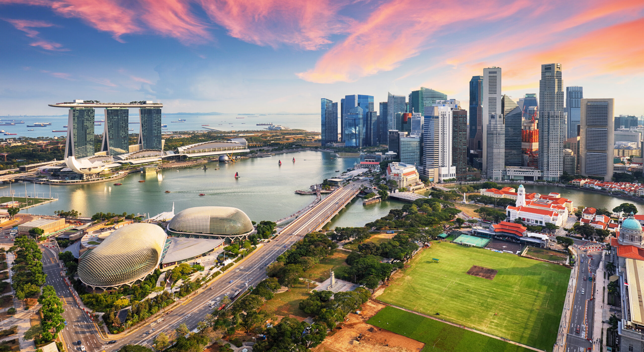 Aerial view of Singapore’s Marina Bay at sunset, showing modern skyscrapers, the Marina Bay Sands hotel, Esplanade domes, roads, and green spaces under a vibrant pink and orange sky.
