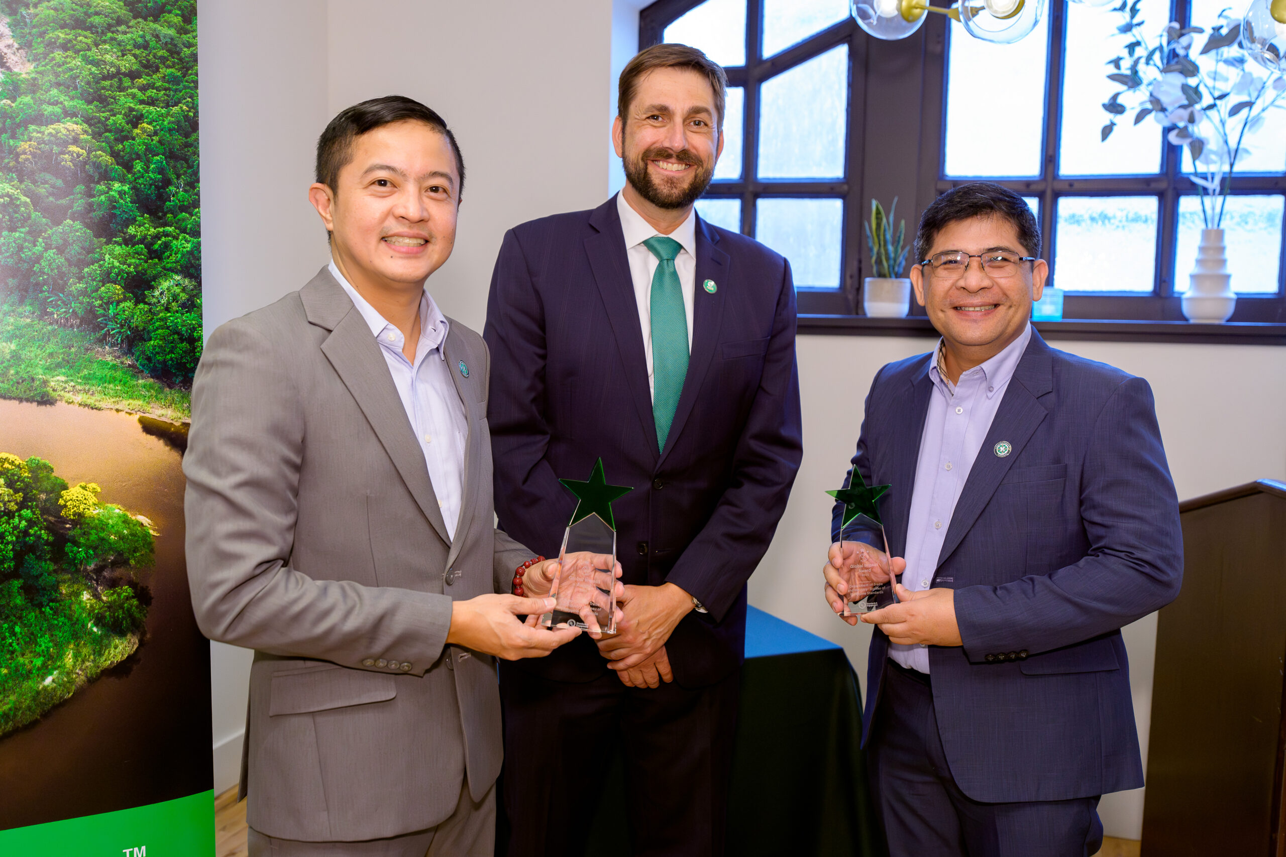 Three men in suits smile at the camera; two hold star-shaped glass awards. They are standing indoors near a window and a banner with a forest image.
