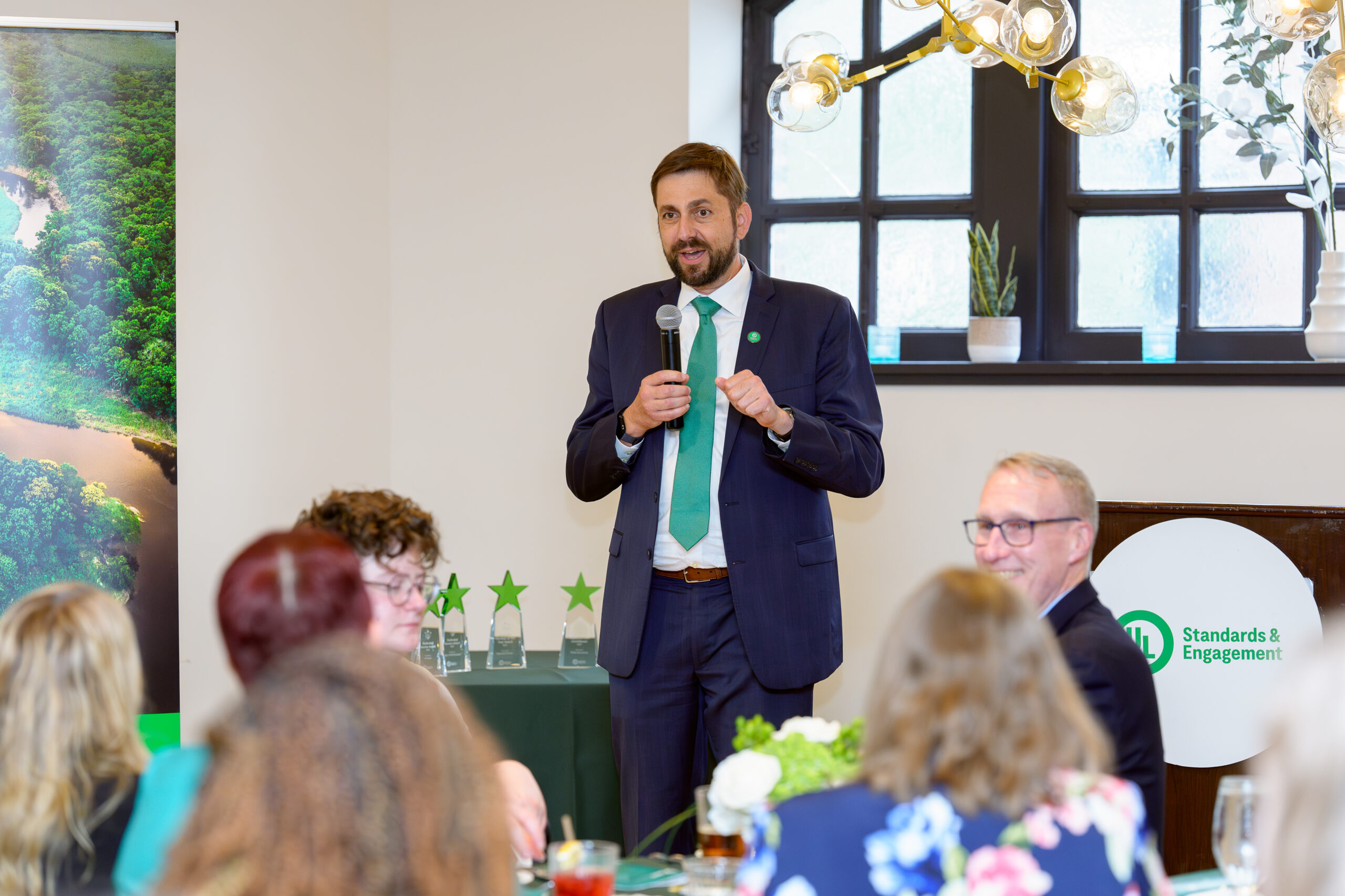 A man in a suit and green tie speaks into a microphone at an indoor event. Several people are seated around tables, and green star-shaped awards are visible on a table behind him. A banner and plants decorate the room.