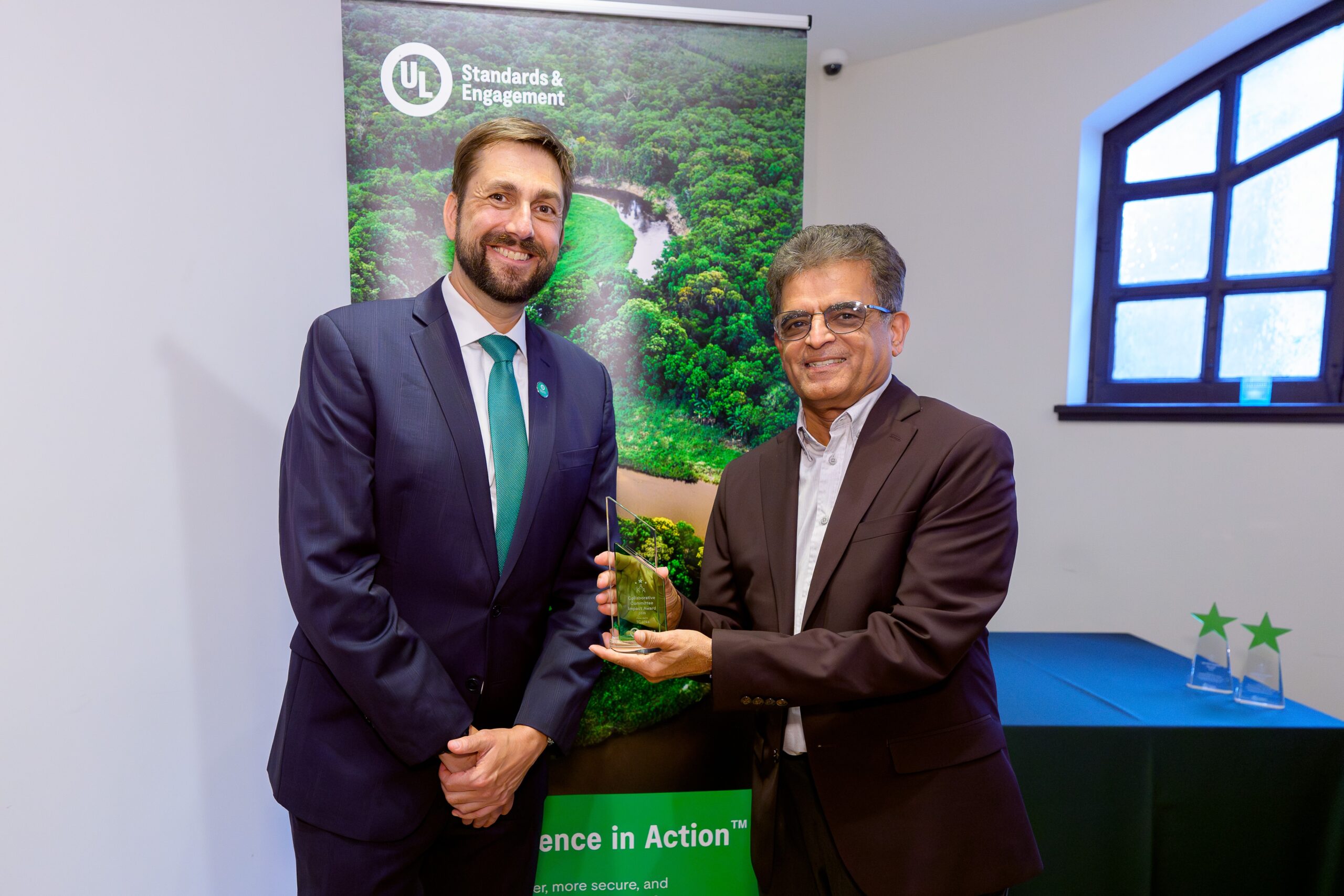 Two men in suits smiling, one handing an award to the other. They are standing indoors in front of a banner with a green landscape and the UL Standards & Engagement logo. A table with trophies is in the background.