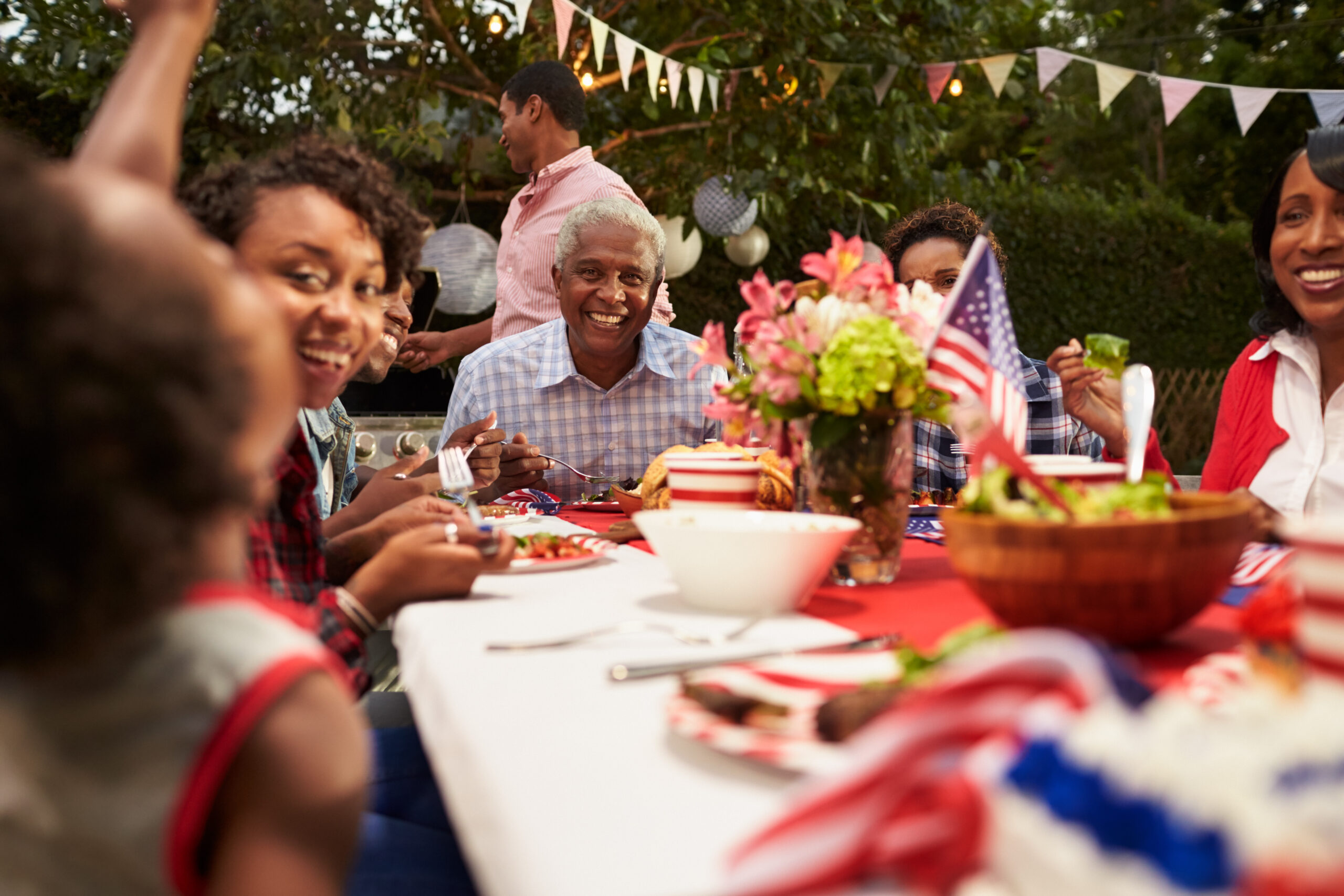 A family gathers around a festive outdoor table decorated with American flags and flowers, sharing a meal and smiling together at a backyard celebration or picnic.