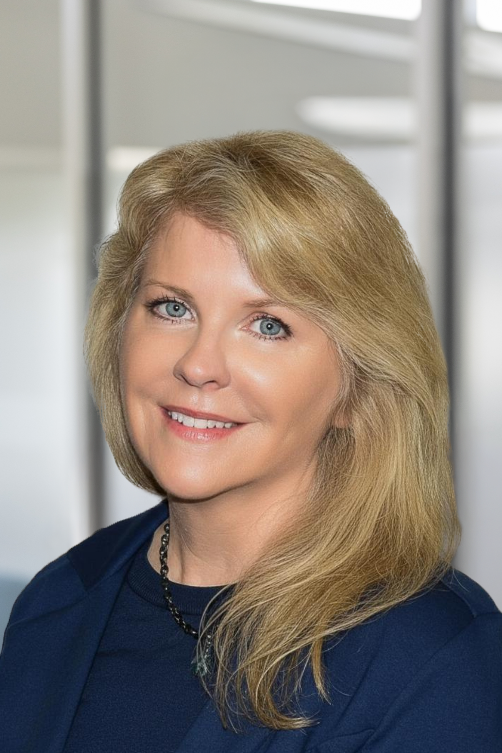 A woman with long blonde hair and blue eyes wearing a navy blue blazer and a necklace, smiling at the camera with a blurred office background.