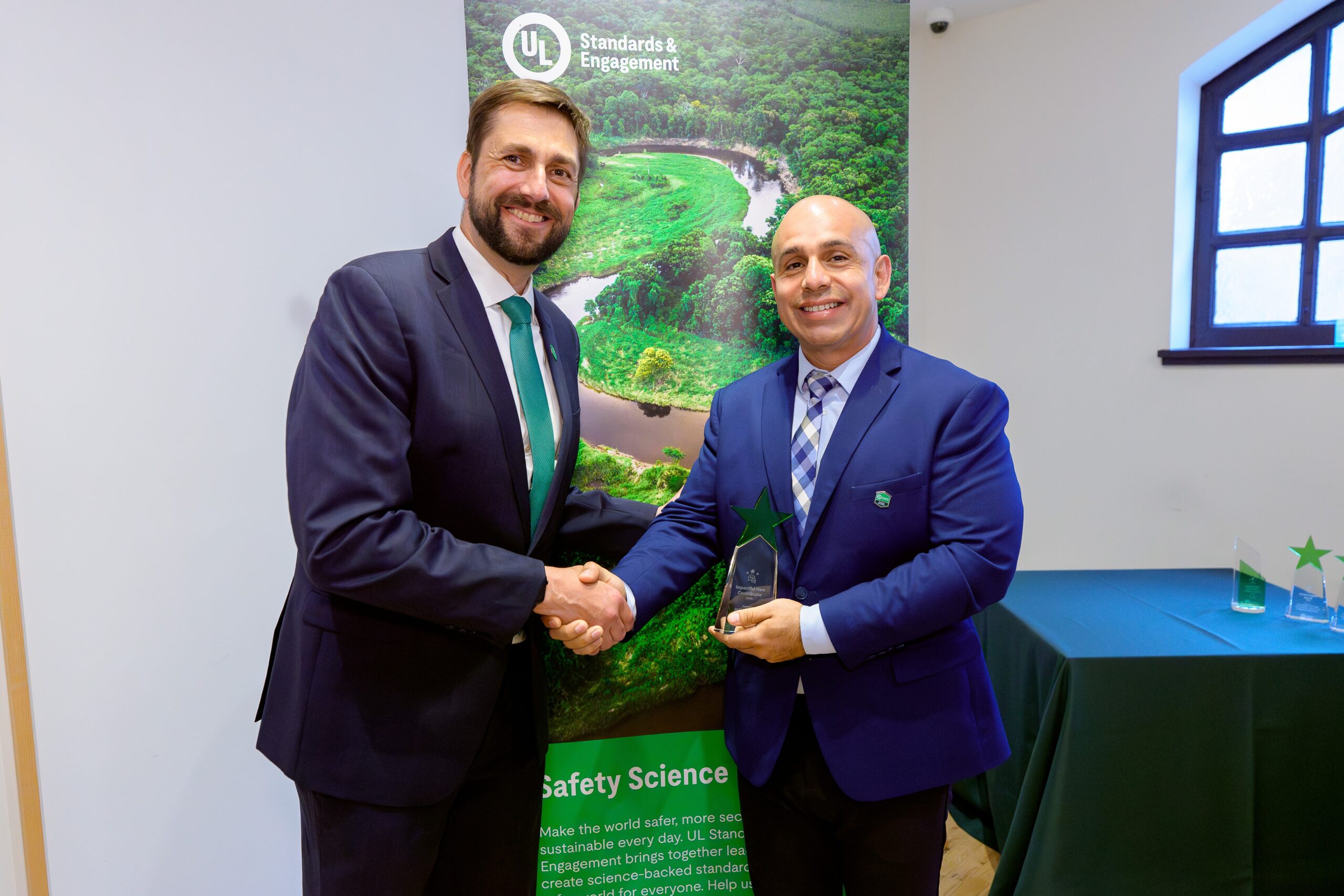 Two men in suits shake hands and smile at an award ceremony. One holds a glass trophy. A UL Standards & Engagement banner and a green-clothed table with other awards are visible in the background.