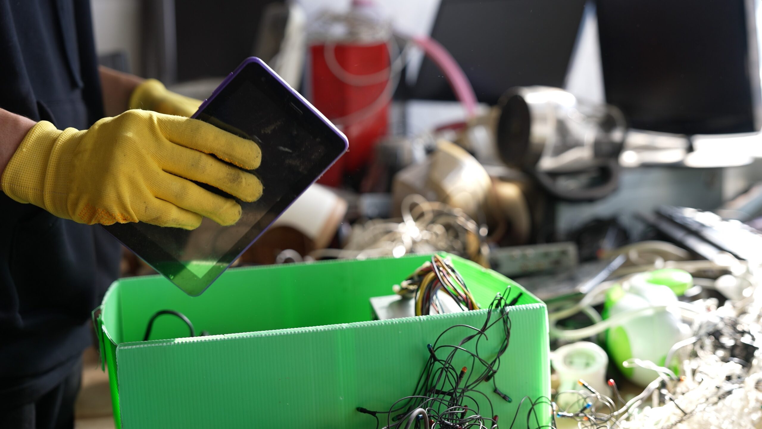A person wearing yellow gloves holds a tablet over a green bin filled with electronic wires and components, suggesting electronic waste recycling. Blurry electronic items and tools are visible in the background.