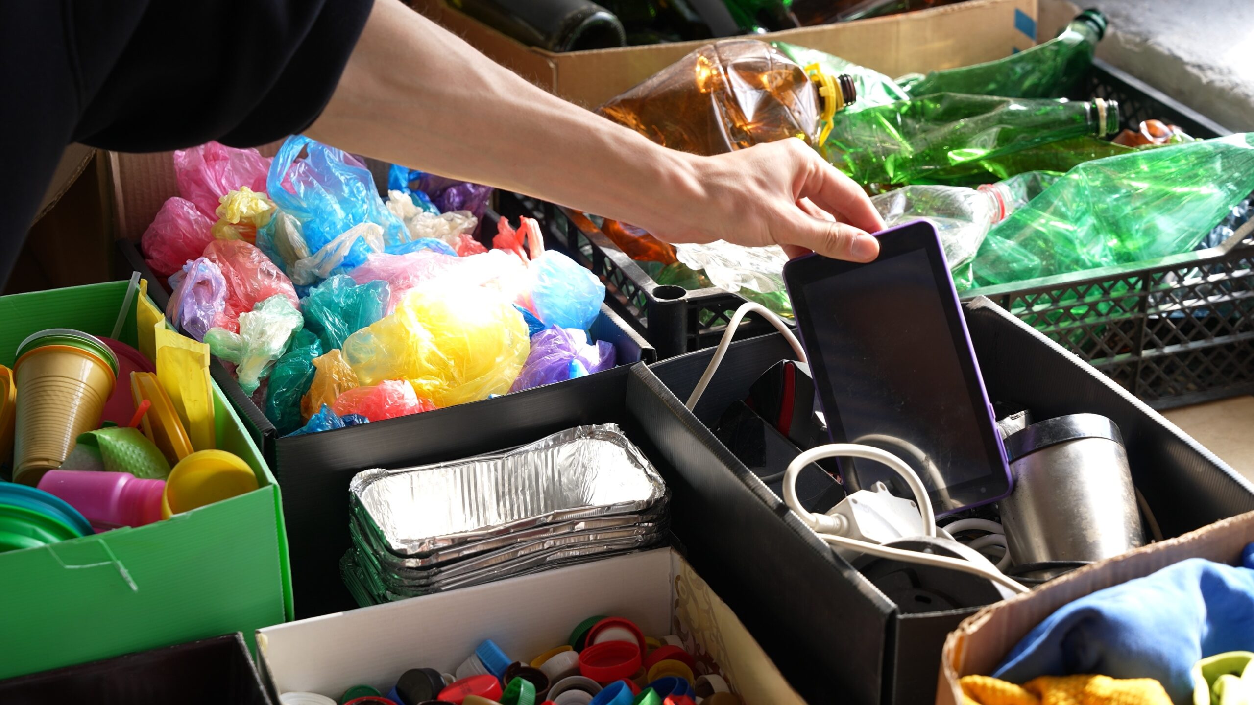 A person’s hand sorts various household waste items for recycling, including plastic bags, glass bottles, aluminum trays, electronics, cables, and colorful plastic lids, all organized in separate boxes.