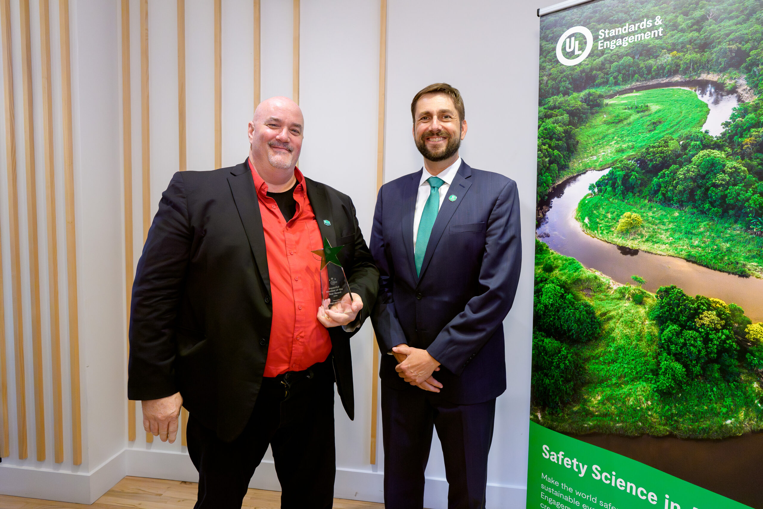 Two men in business attire stand smiling indoors; one holds a small award. Next to them is a banner with a nature scene and the text “UL Standards & Engagement” and “Safety Science in Action.”.
