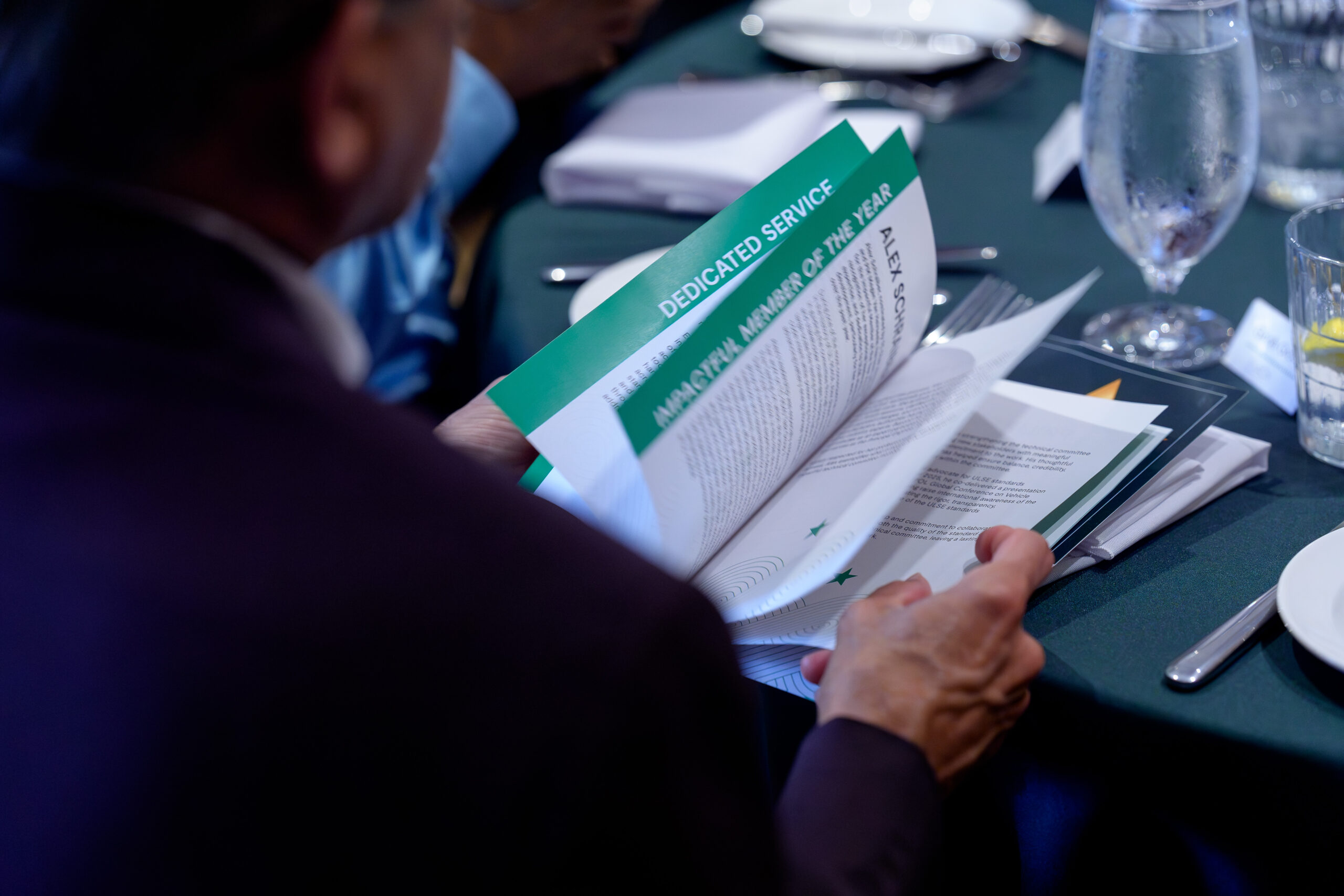 A person sits at a table reading a booklet titled "DEDICATED SERVICE MUNICIPAL MEMBER OF THE YEAR," with water glasses, plates, and silverware visible nearby. The scene suggests a formal event or ceremony.