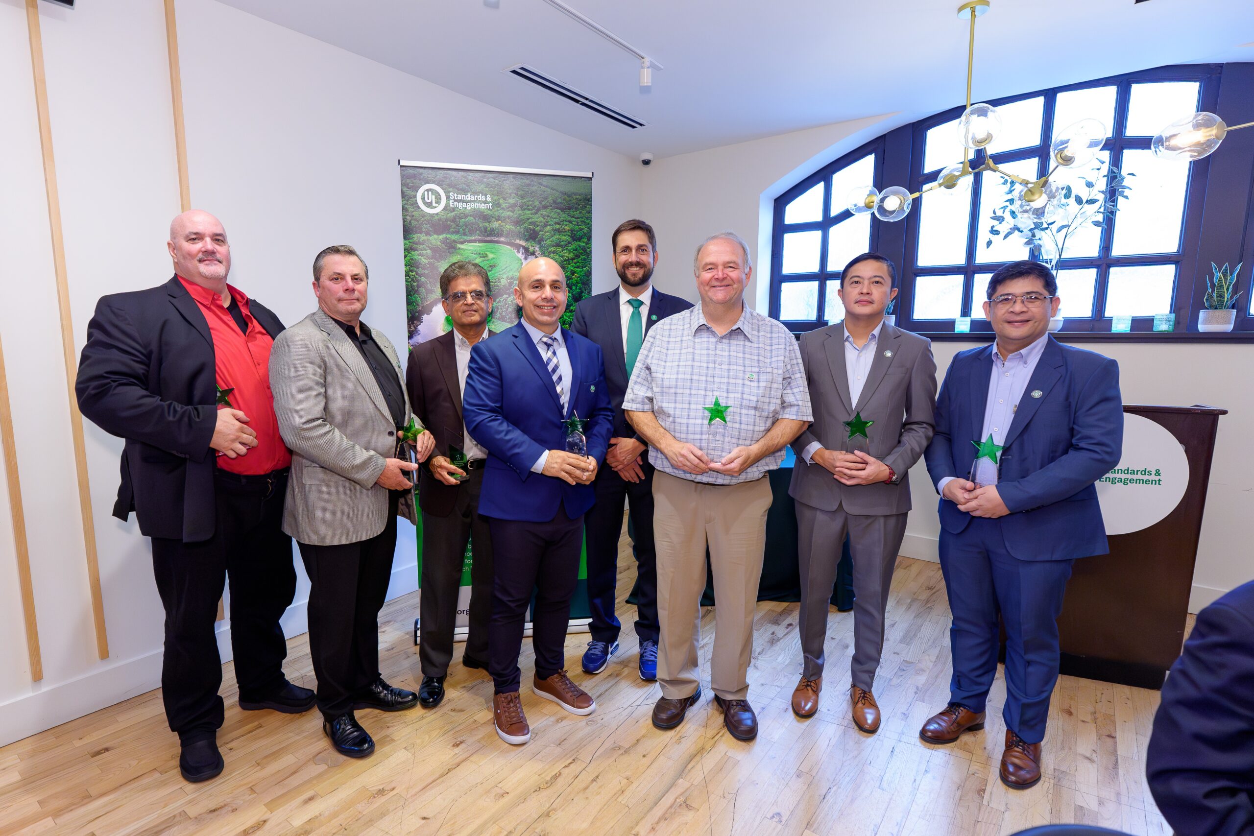 Eight men in business attire stand in a well-lit room, some holding green star-shaped awards, posing for a group photo in front of a sustainability-themed banner and a podium.