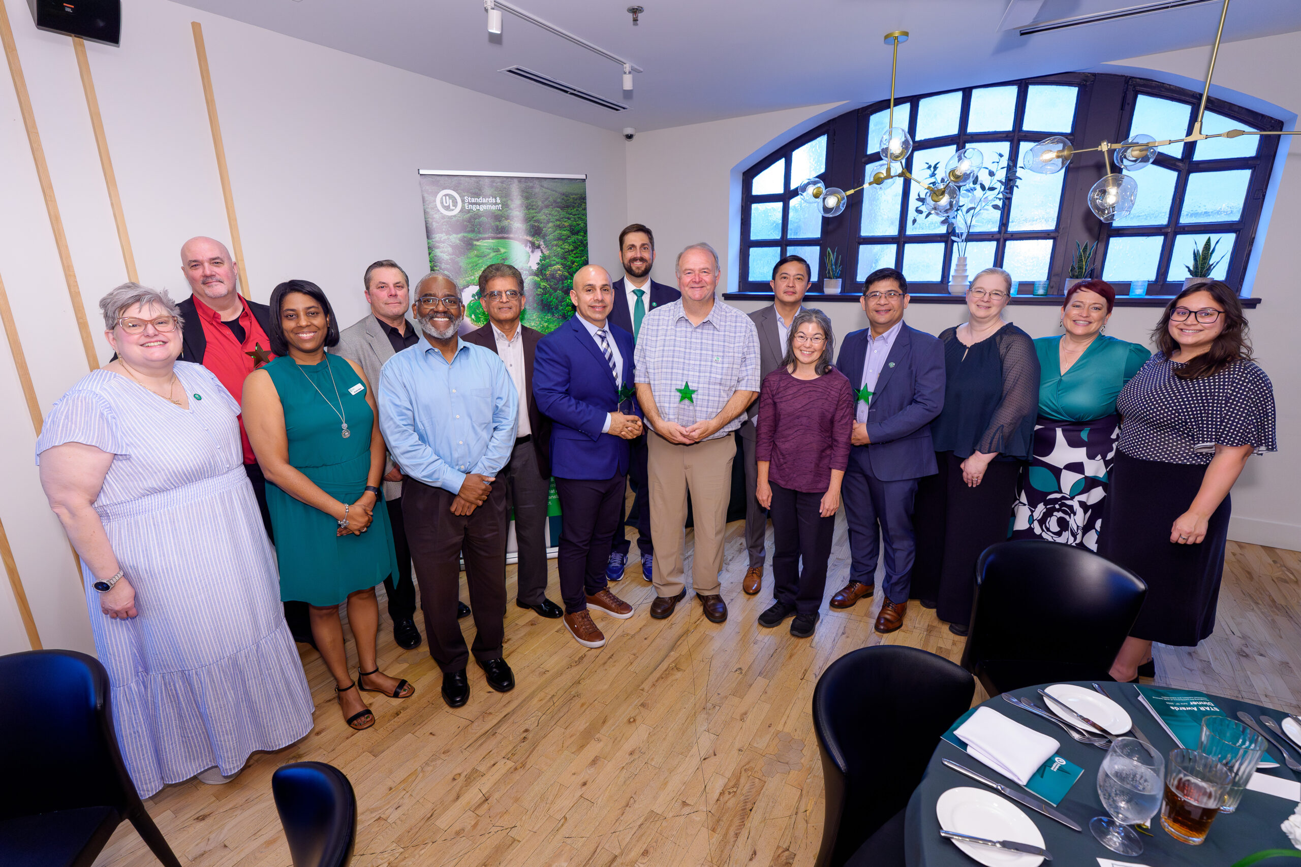 A group of fifteen adults, both men and women, stand together smiling in a well-lit room with wooden floors and large windows. They are dressed in business attire, some holding green star-shaped awards.