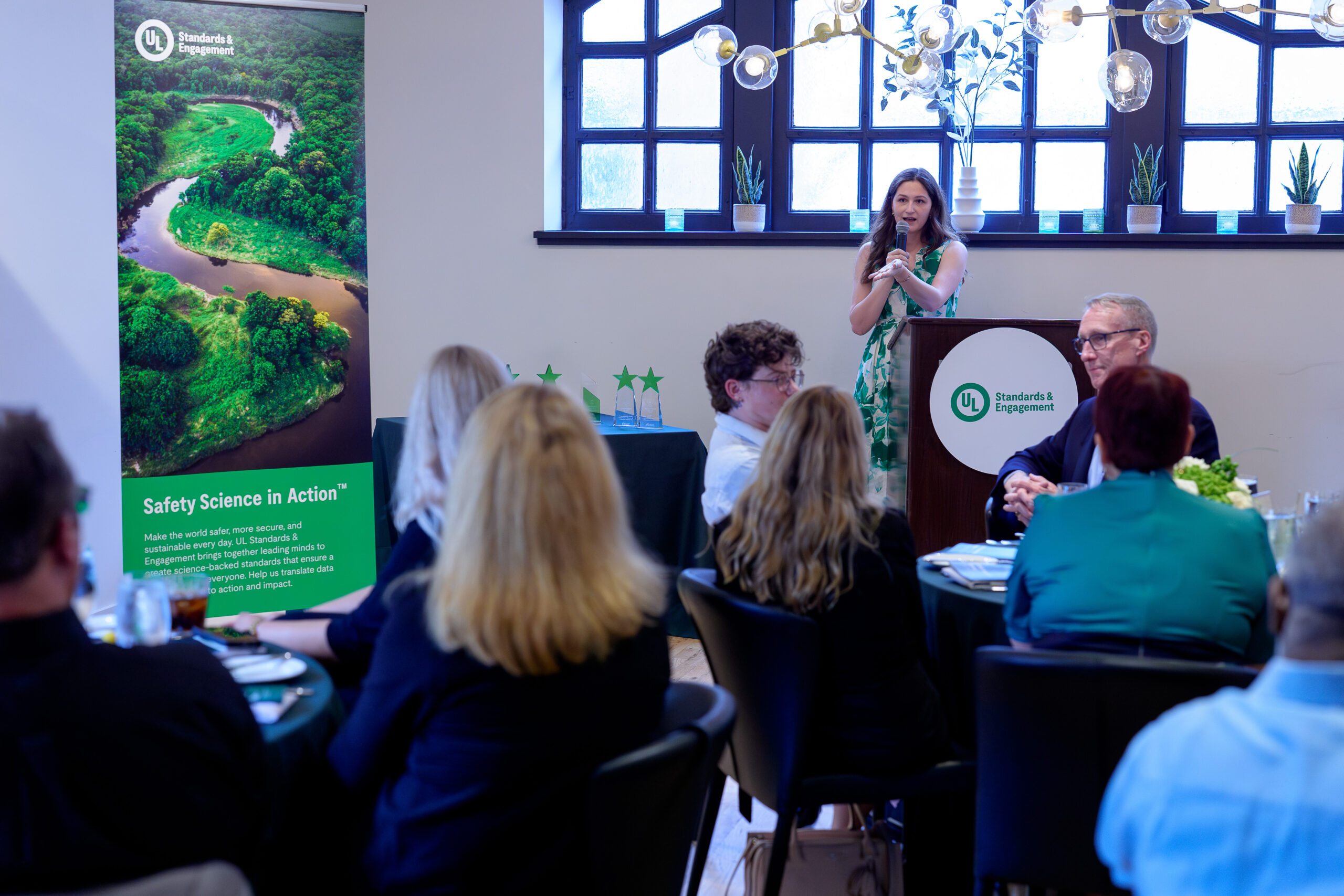 A woman speaks at a podium to a seated audience in a well-lit room; a banner on the left displays a river and the words "Safety Science in Action.