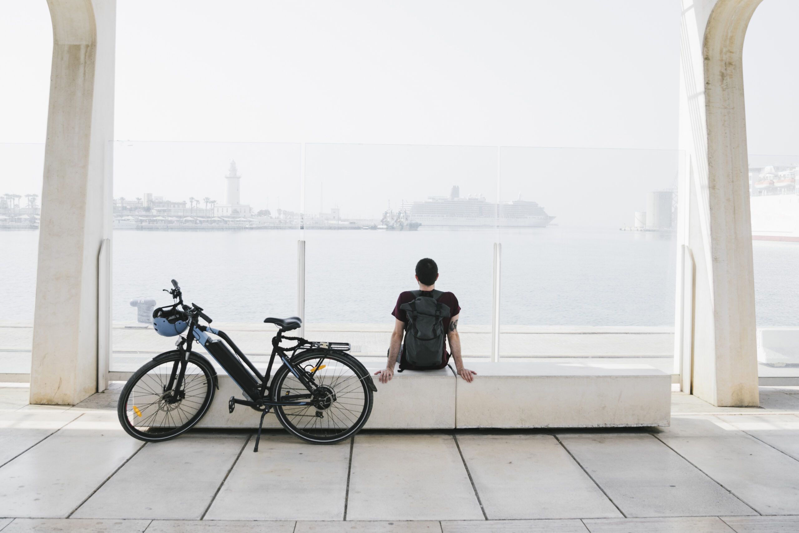 A person with a backpack sits on a bench facing the water, with a bicycle parked beside them. In the distance, a large ship and a lighthouse are visible through the mist at a harbor.