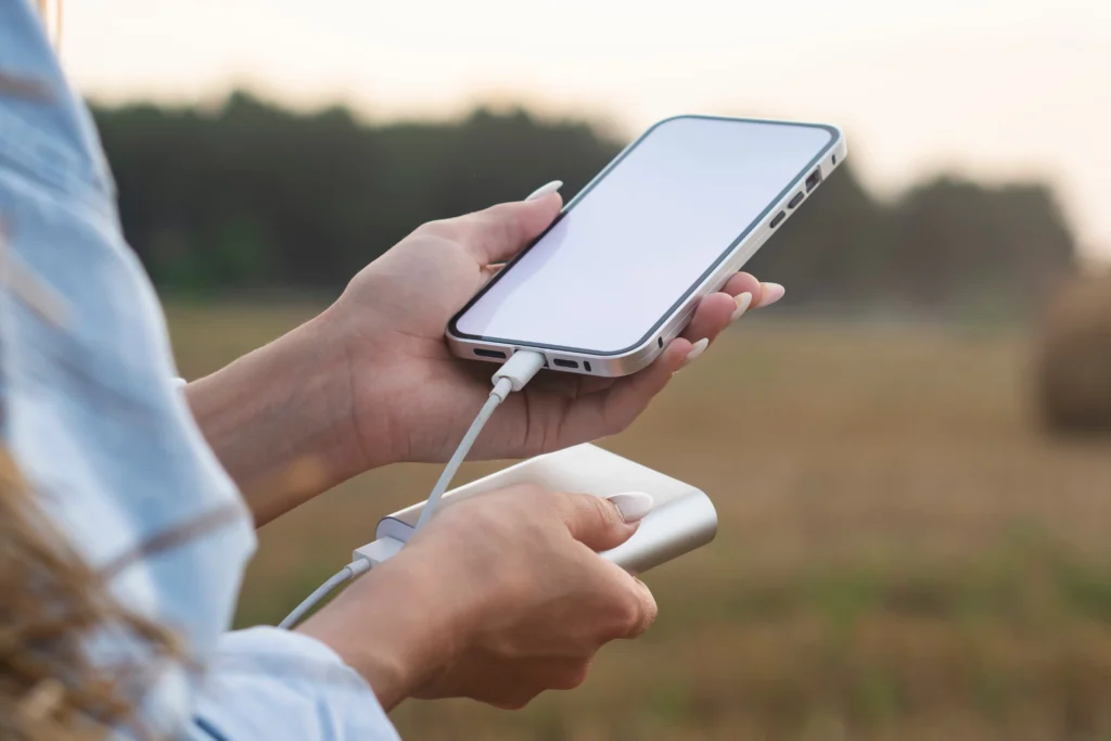 A person holds a smartphone connected to a portable power bank, charging the device outdoors with a blurred natural background.