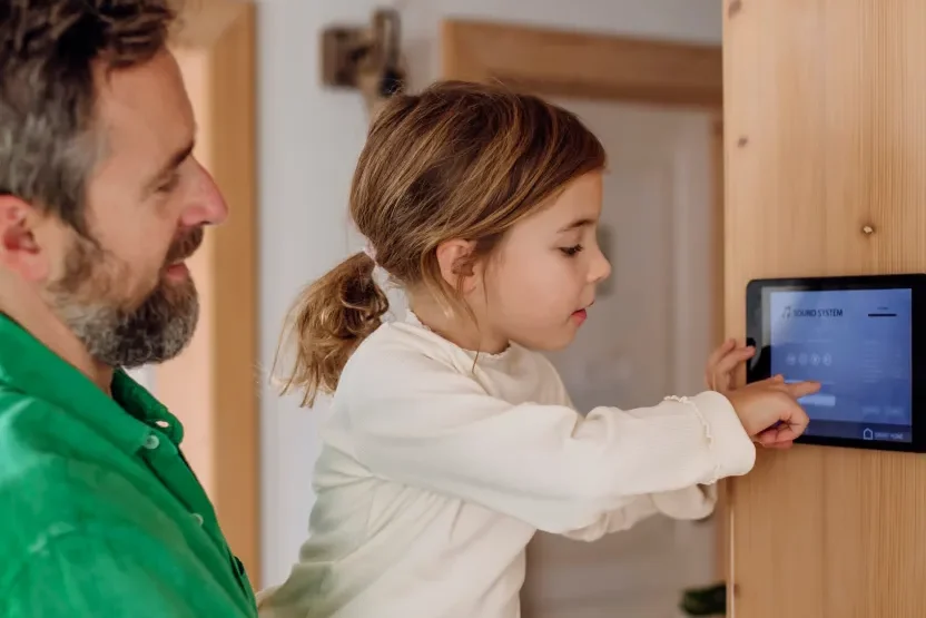 A man holds a young girl as she touches a smart home control panel mounted on a wooden wall, both focused on the screen. The setting appears to be a modern home interior.